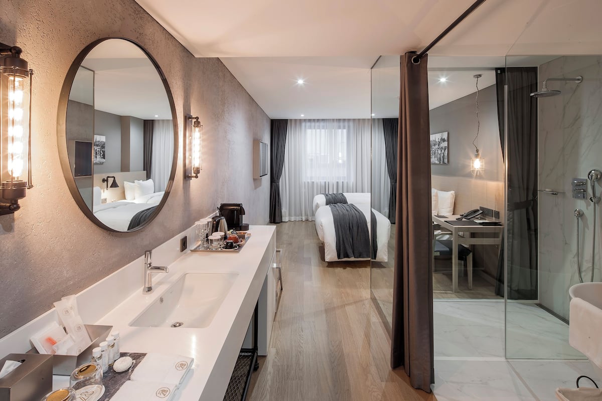 A modern hotel room with a large mirror, a long vanity with a sink, a bed, a desk, and a glass-enclosed shower area.
