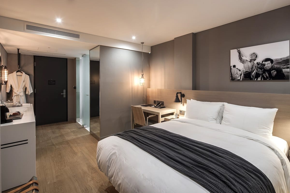 A modern hotel room with a large bed, a desk, a chair, a wardrobe, and a mirror.