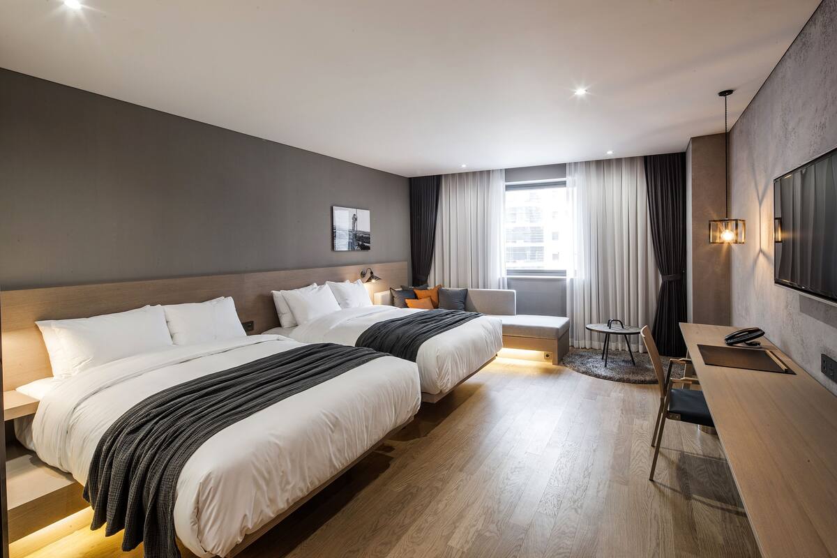 A modern hotel room with two beds, a desk, a chair, a TV, and a window with curtains.