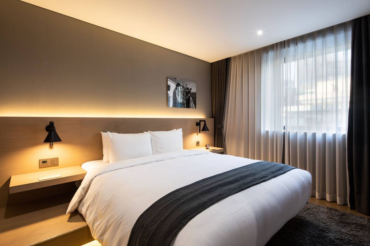 A modern hotel room with a large bed, bedside tables, a wall-mounted light, and a window with curtains.