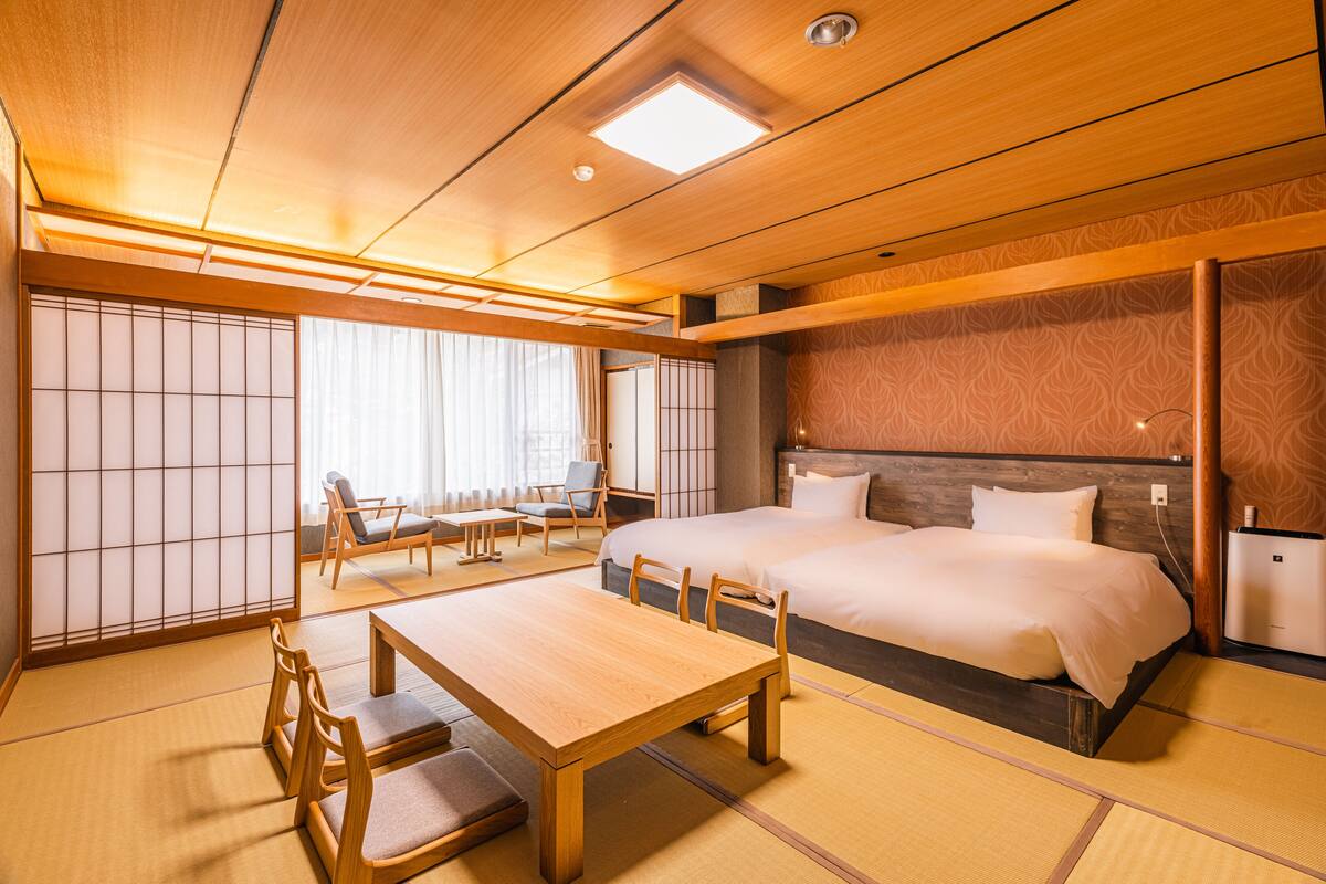Una habitación japonesa tradicional con suelo de tatami, una mesa de madera baja con sillas, una cama con cabecero de madera y un panel corredizo shoji.