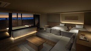 Une chambre d’hôtel moderne dotée d’une grande fenêtre donnant sur un paysage urbain au coucher du soleil, d’un coin salon confortable et d’un lit soigneusement fait.