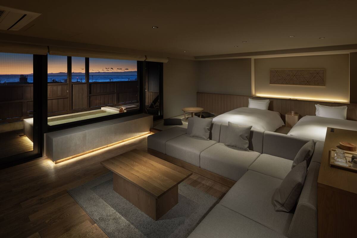 Un hotel moderno con grandes ventanales que dan a una vista urbana al atardecer, un rincón de descanso acogedor y una cama bien ordenada.