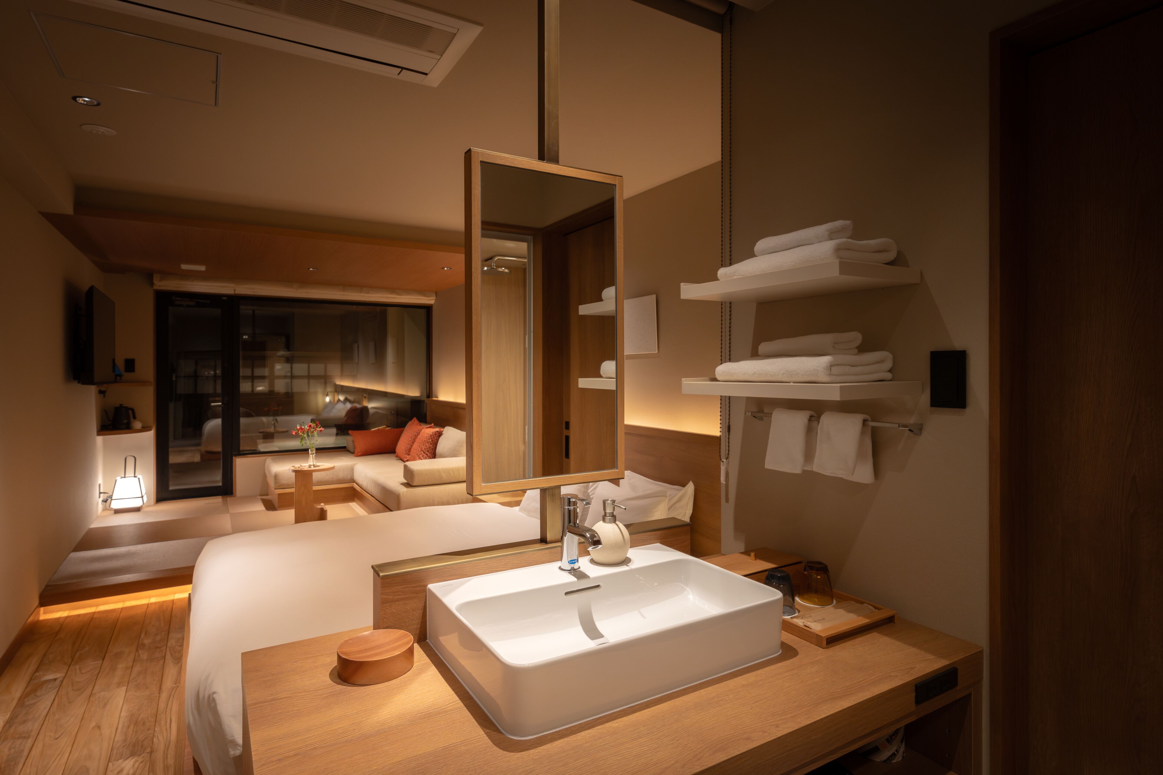 A modern hotel bathroom with a large mirror, a sink, and a wooden vanity. There is a bed with a view of the room and a small table with a lamp.