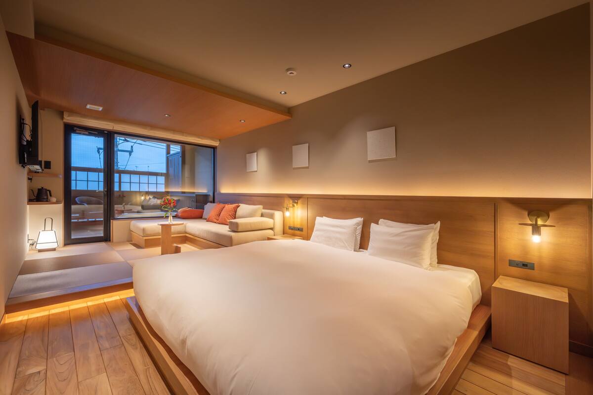 Habitación de hotel moderna con una cama grande, una zona de estar con sofá y sillones, una mesita y vistas al exterior a través de una puerta corredera.