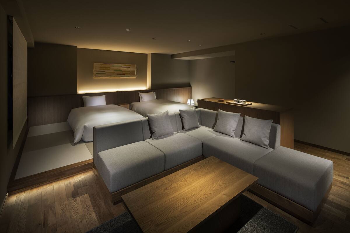 Una habitación de hotel moderna con sofá modular, dos camas, una mesita de madera y una lámpara de pared.