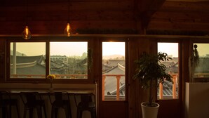 Quarto com paredes de madeira, uma janela grande com vista para arquitetura tradicional, uma planta em vaso e luminárias pendentes.