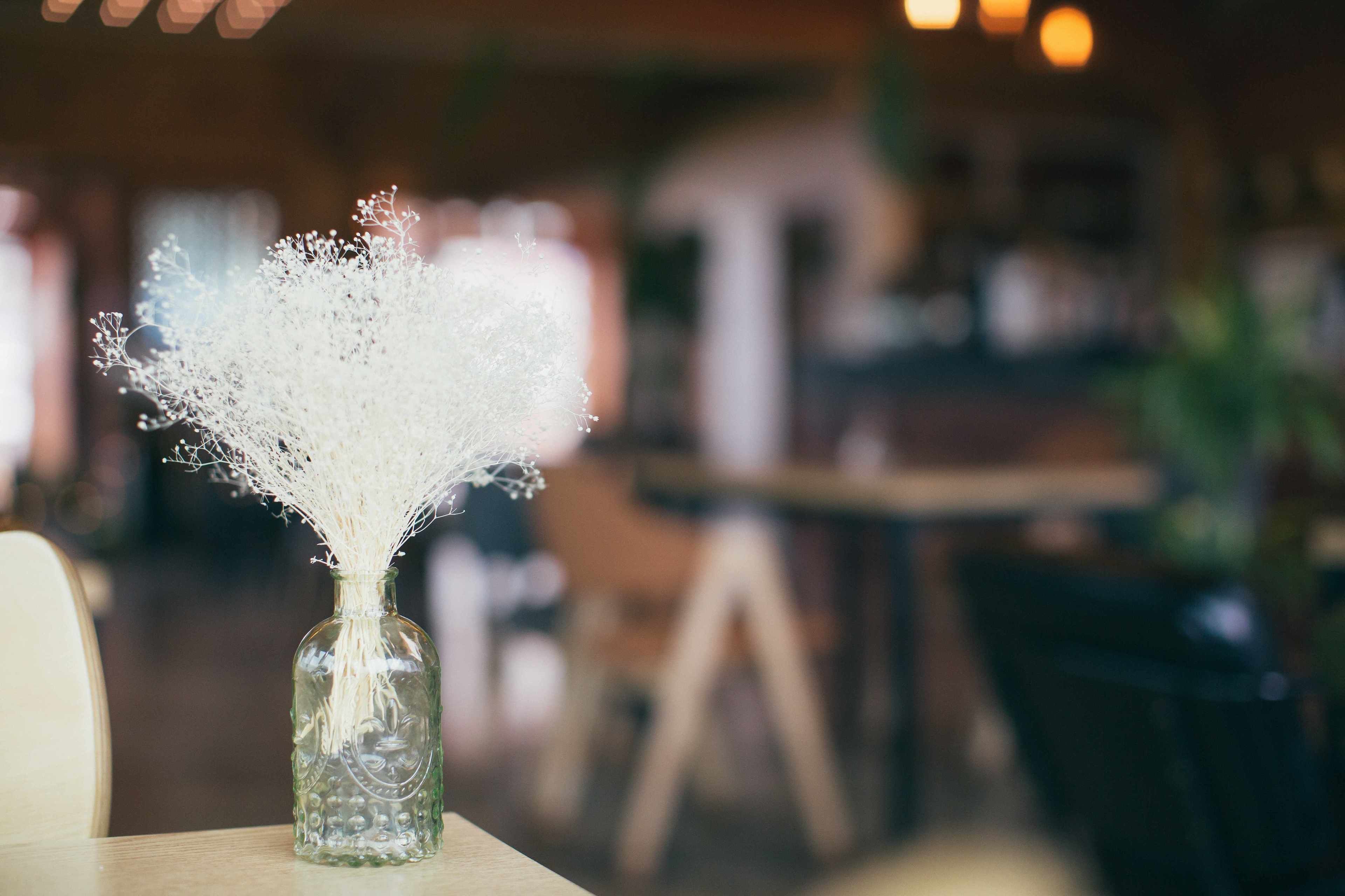 Eine transparente Glasvase mit einer weißen, filigranen Blumenkomposition auf einem Holztisch in einem Restaurant.