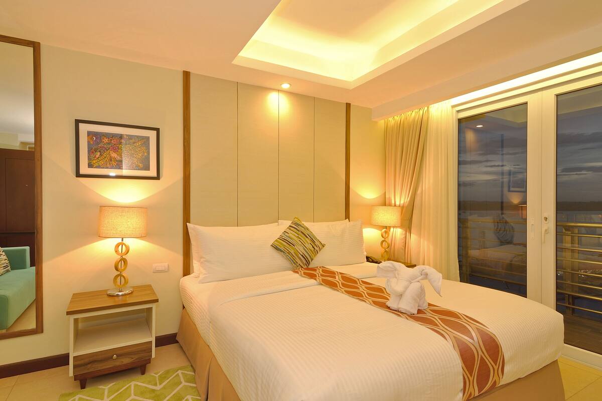 Quarto de hotel com uma cama grande, duas luminárias de cabeceira, uma obra de arte emoldurada na parede e vista pela porta de vidro.