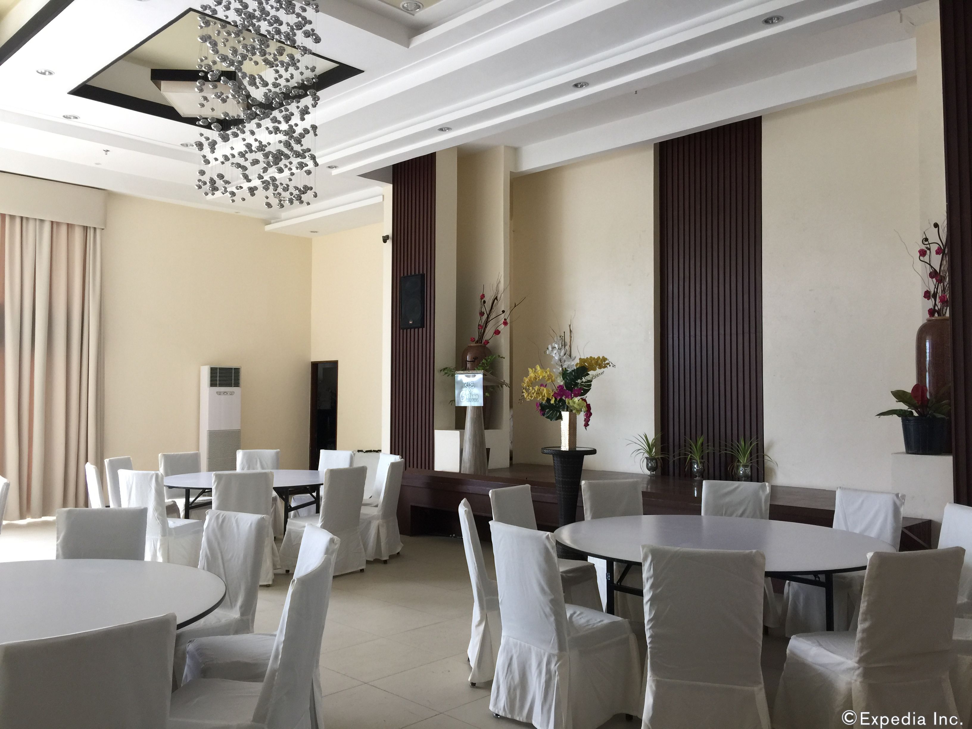 A hotel conference room with round tables and white chairs, a modern chandelier, and decorative plants.