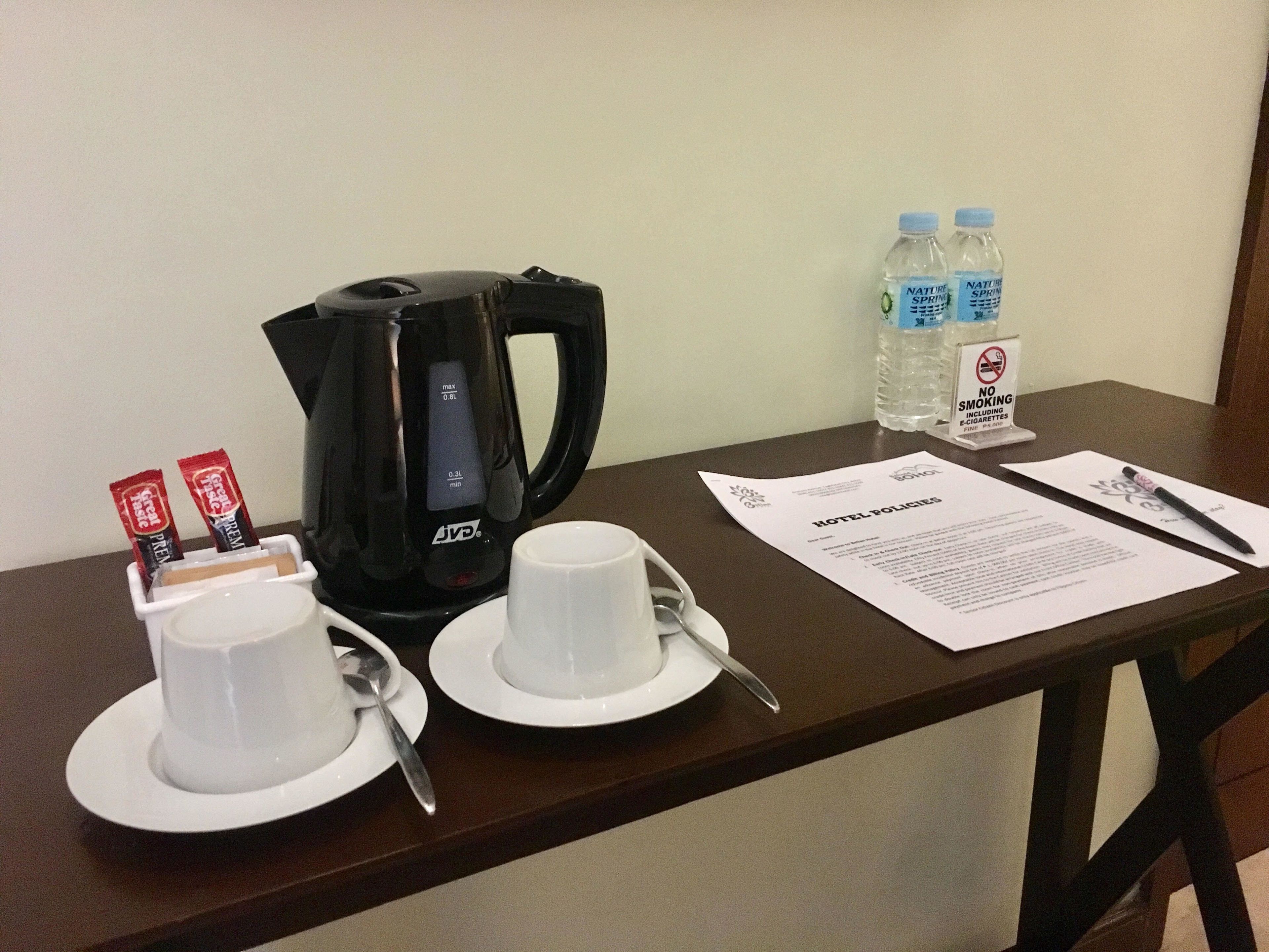 Een hotelkamer tafel met een zwarte koffiezetapparaat, twee witte bekers, een lepel, een waterfles en een bordje ‘Niet roken’.