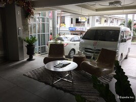Un hall d’hôtel avec un comptoir d’accueil, un espace salon comprenant deux fauteuils et une table, une plante en pot et un fourgon stationné à l’extérieur.