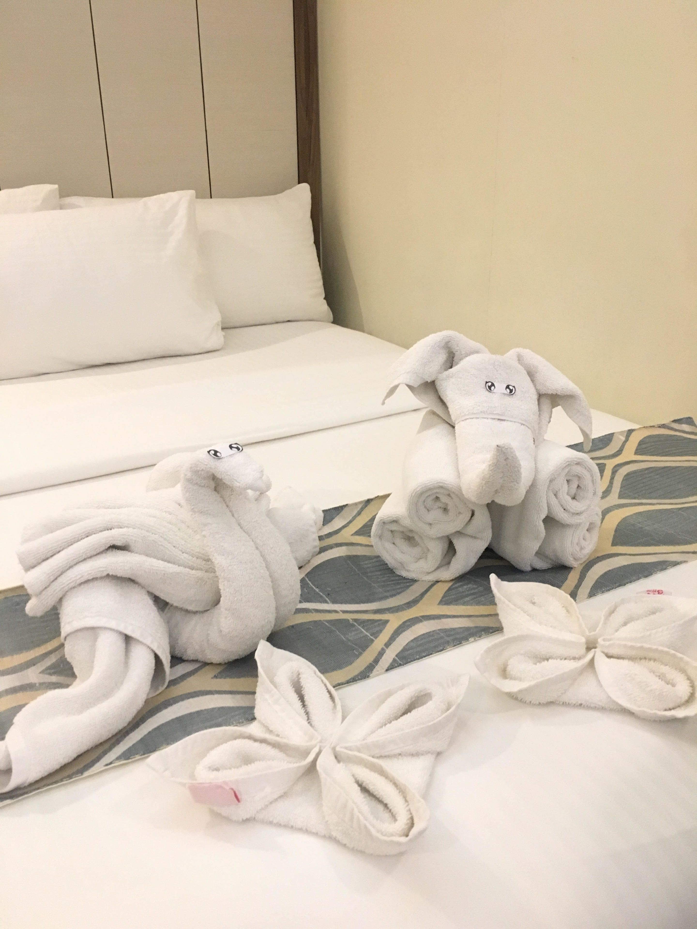 Un letto rifatto con lenzuola bianche, un peluche a forma di coniglio e asciugamani arrotolati disposti su un cuscino decorato.