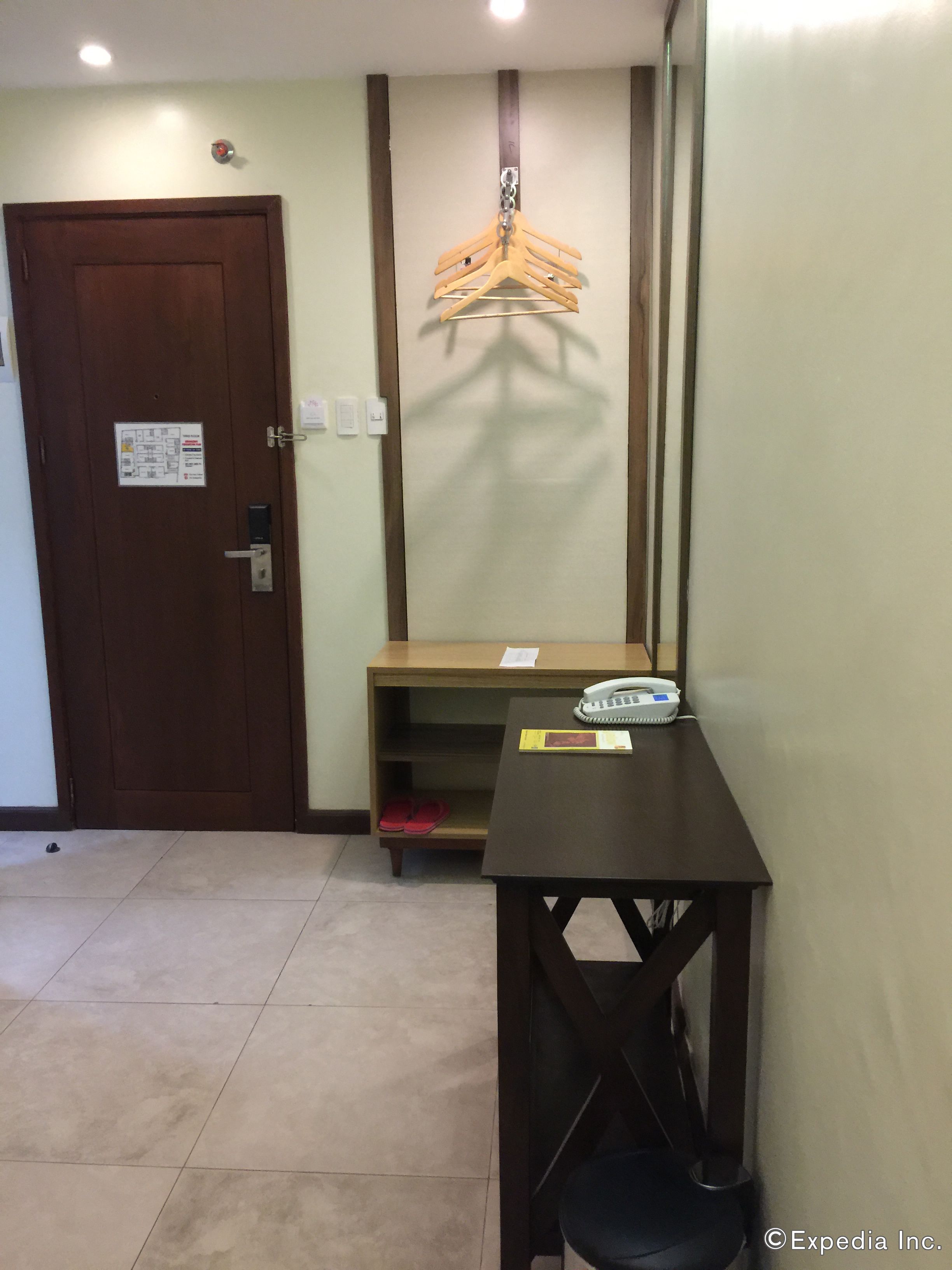 A hotel room with a wooden door, a coat rack, a small table with a telephone, and a pair of slippers on the floor.