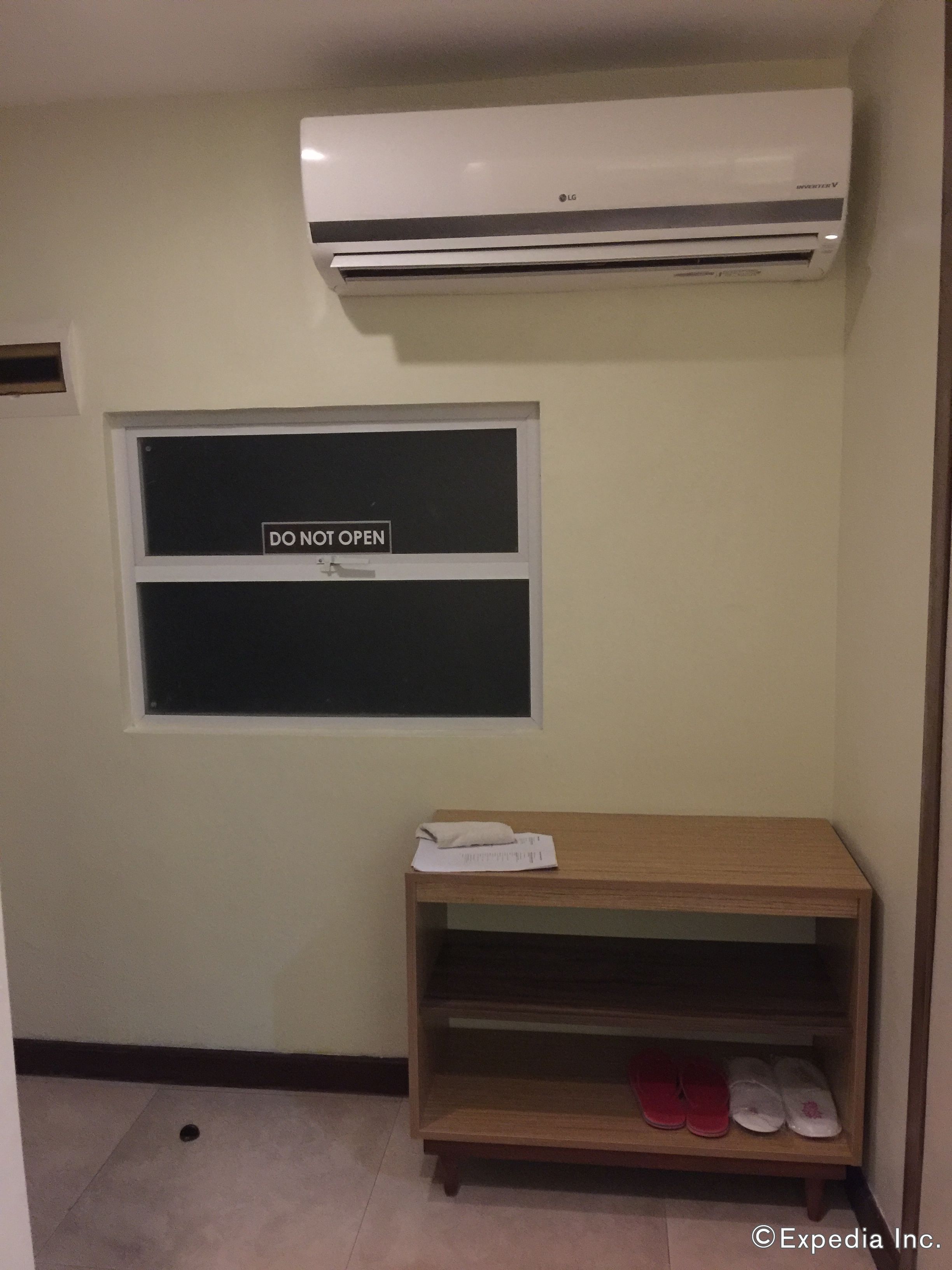 Una habitación con aire acondicionado blanco, una ventana que indica 'NO ABRIR' y un estante de madera con zapatillas.