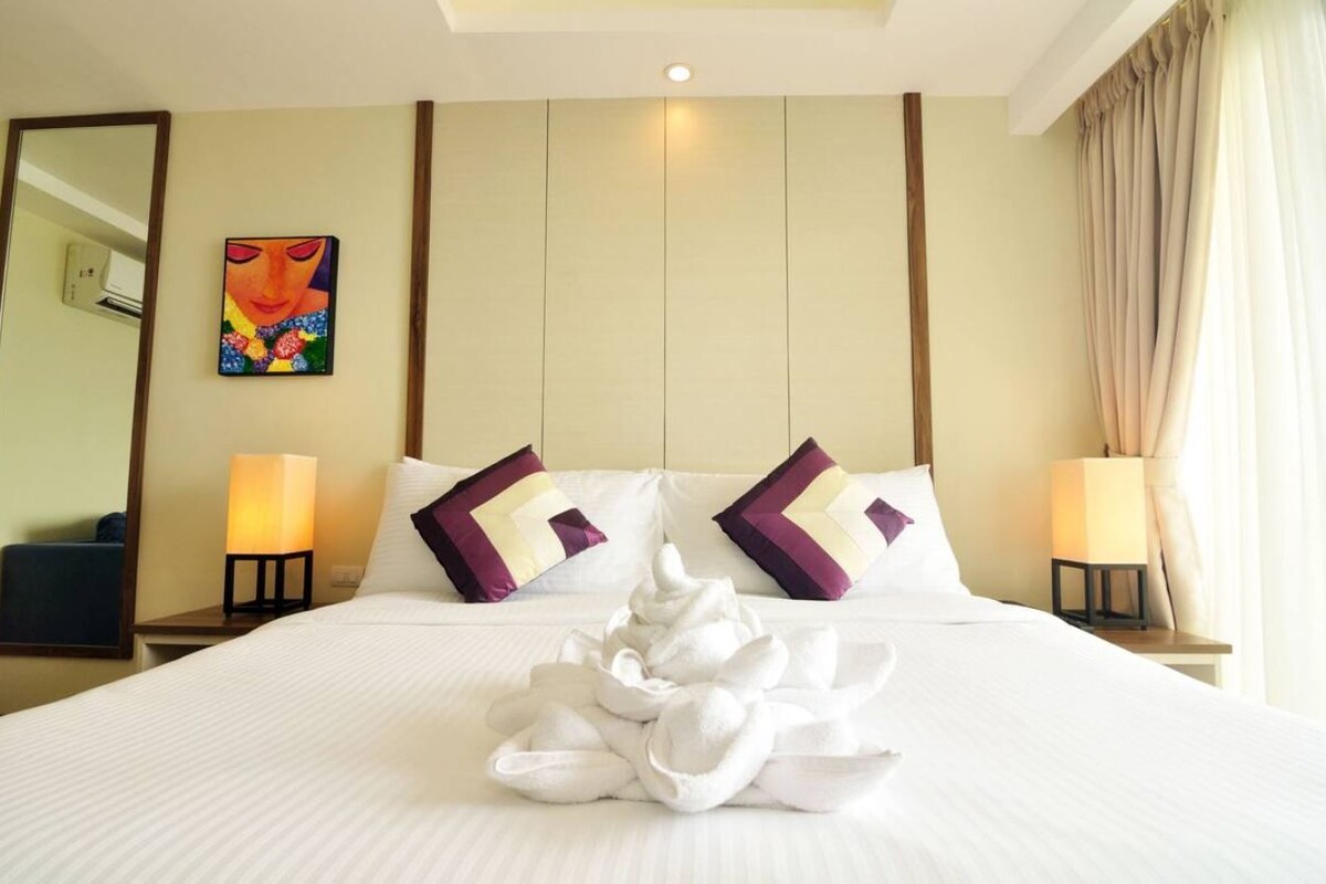 A hotel room with a neatly made bed, two bedside lamps, a painting on the wall, and a large window with curtains.