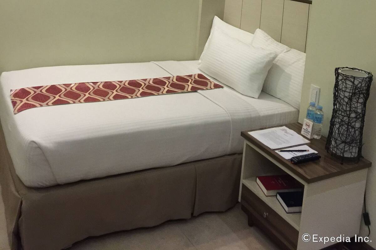 Uma cama bem arrumada com um edredom estampado, uma mesinha de cabeceira com uma luminária e uma pequena prateleira com livros e uma garrafa.