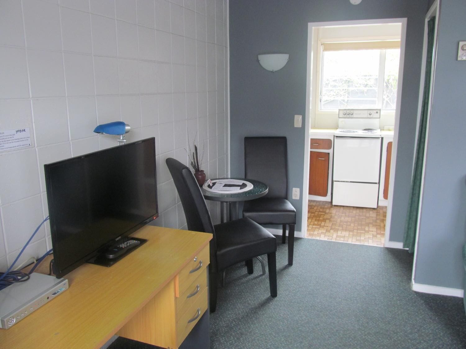 A compact room with a desk, chair, and a small table. A television is on the desk, and a window provides natural light.