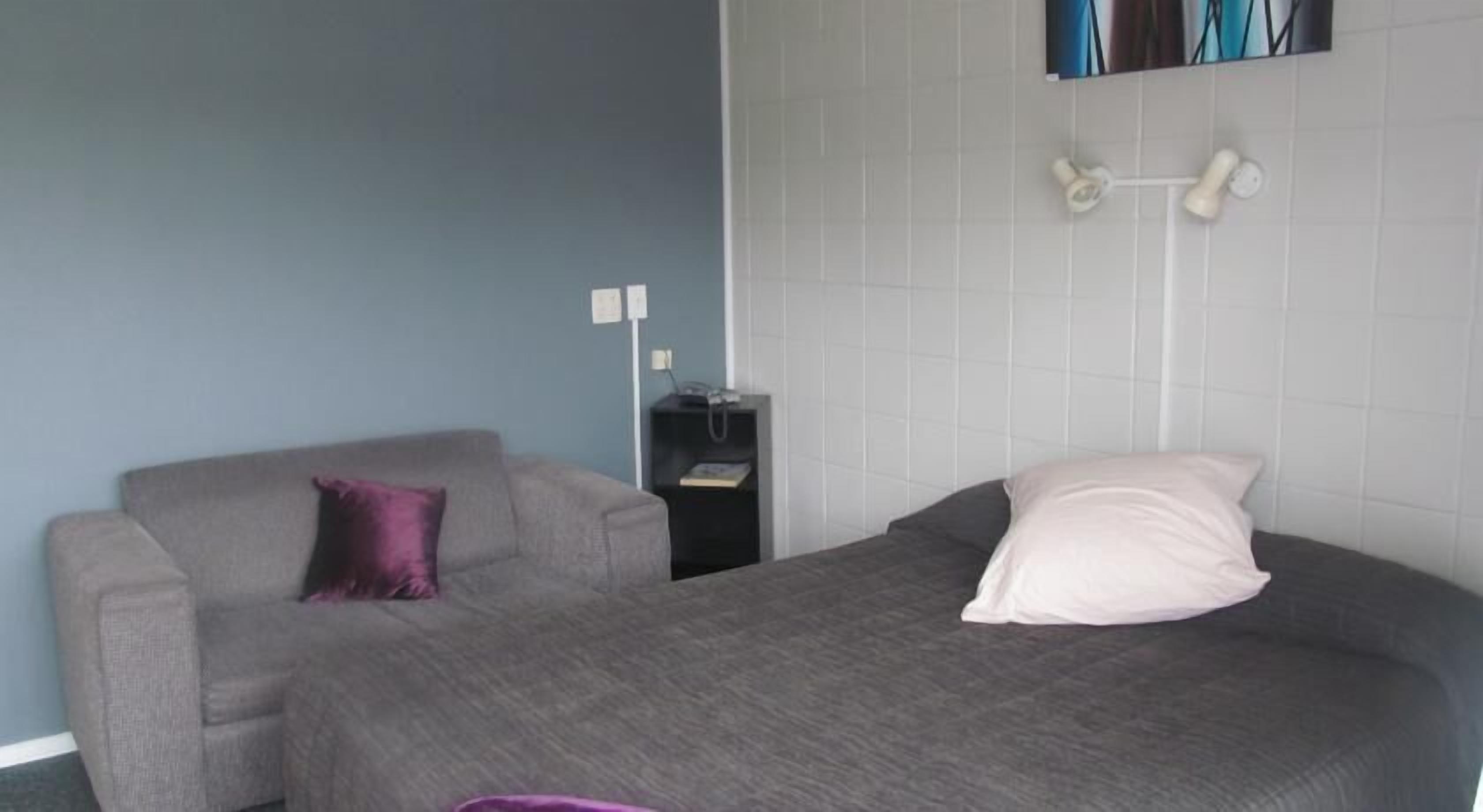 A small, modern room with a grey sofa and a single bed. There is a nightstand with a lamp and a small shelf with books.