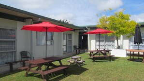A patio area with picnic tables and red umbrellas, a small tree, and a building with windows and a door. - Parkview Motel (Dargaville)