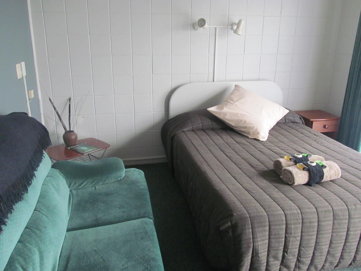 A hotel room with a bed, a green sofa, a small table with a vase, and a nightstand.