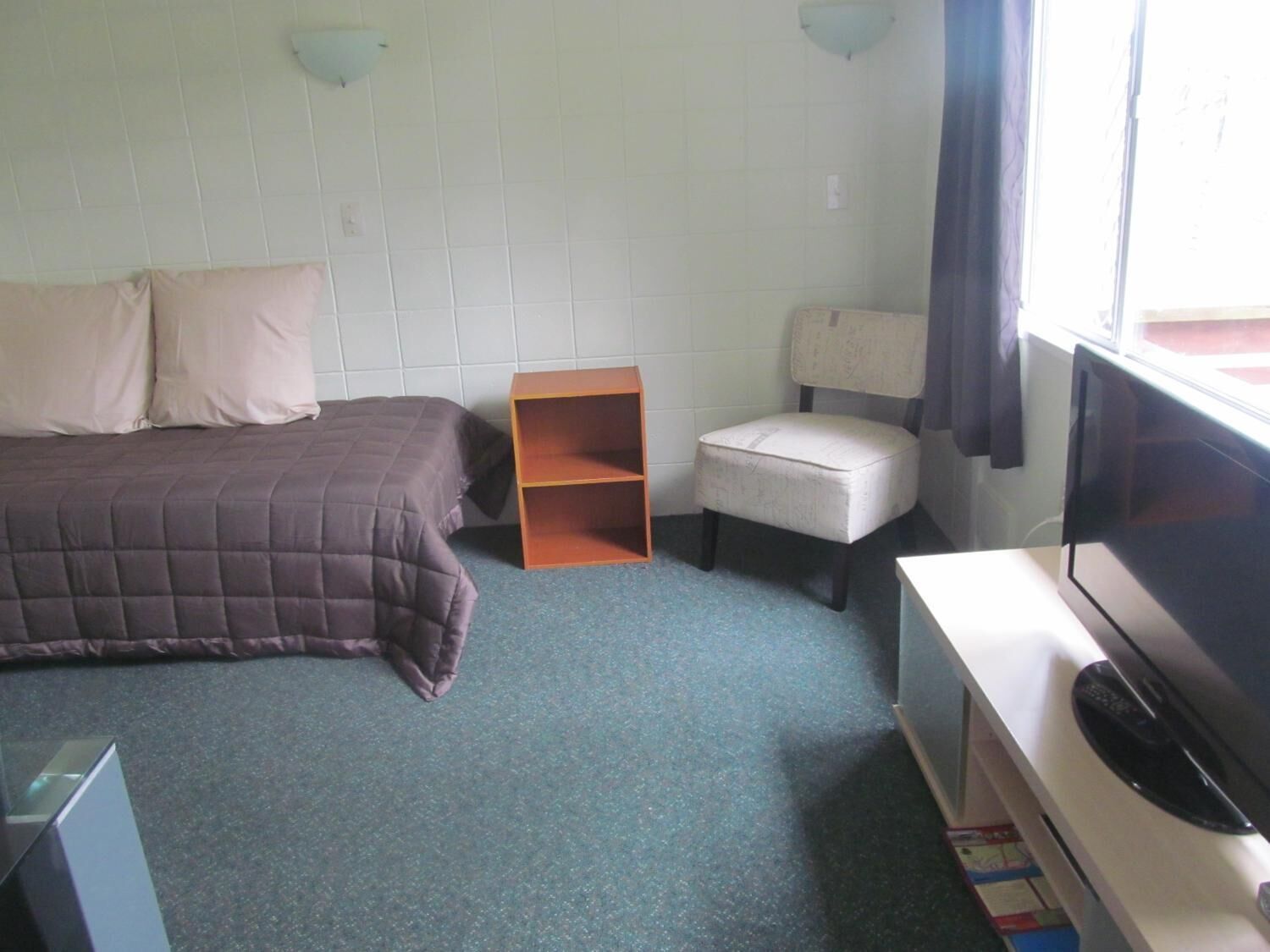 A bedroom with a bed, a chair, a TV, and a small wooden shelf.