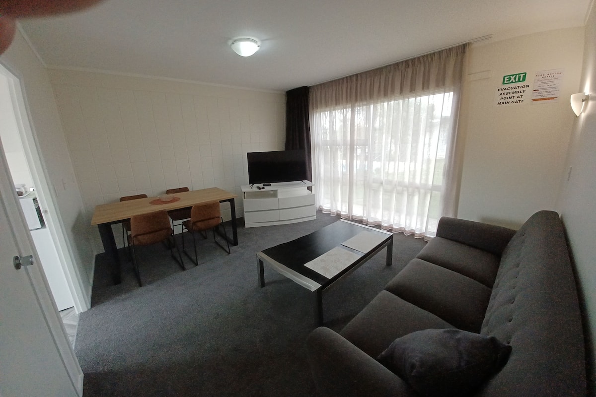A living room with a sofa, a coffee table, a TV on a stand, a dining table with chairs, and a large window with curtains.