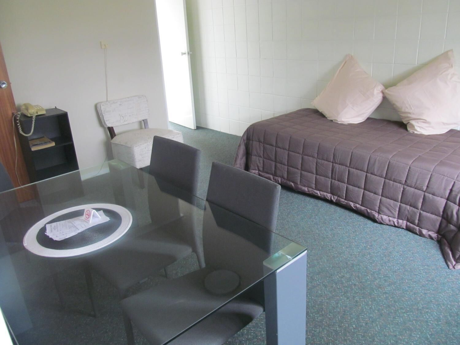 A hotel room with a glass table, two chairs, a bed with a purple blanket, and a white wall.