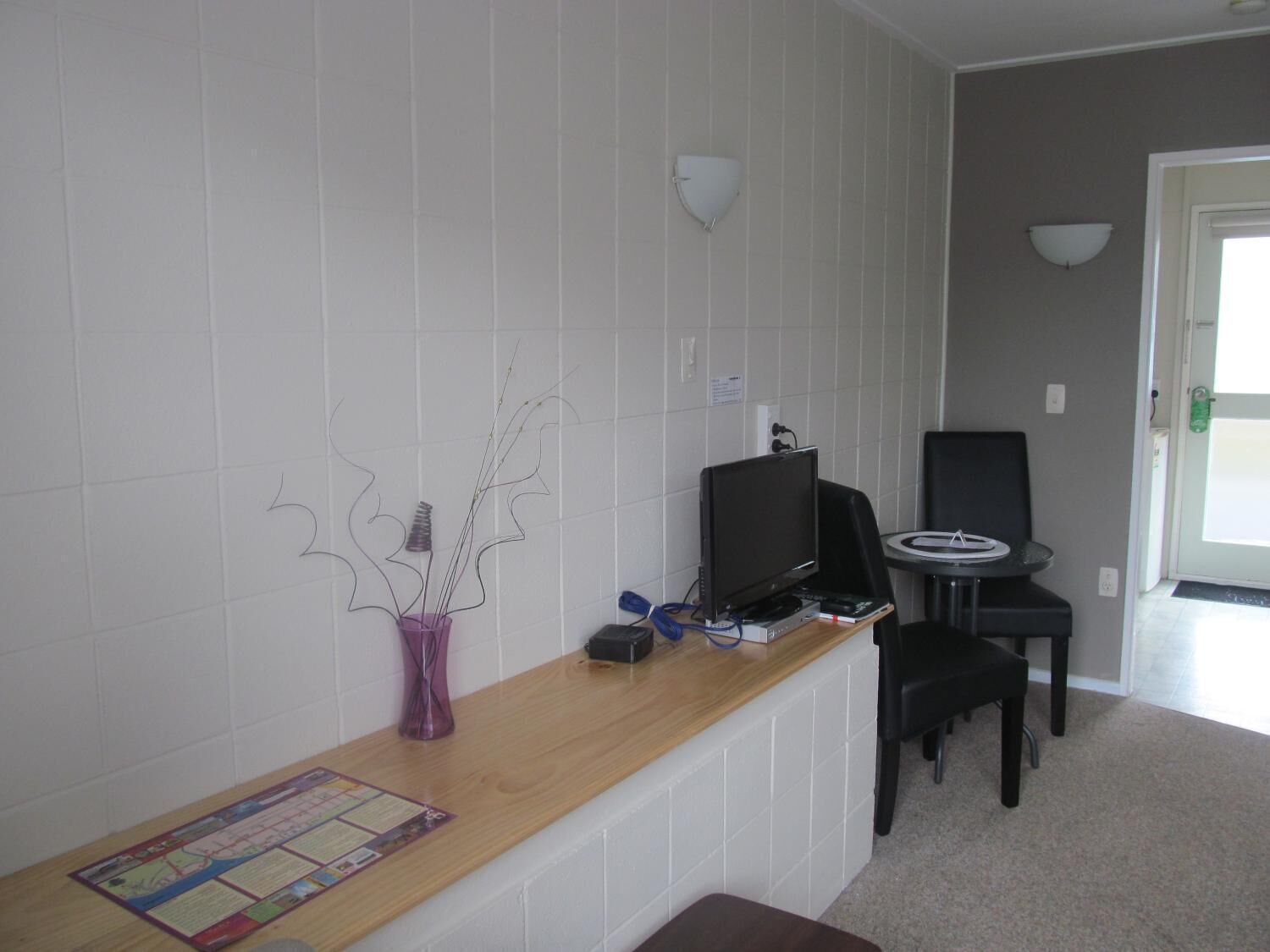 Una habitación pequeña y limpia con un mostrador de madera, una silla negra, una mesita y un jarrón morado con ramas.
