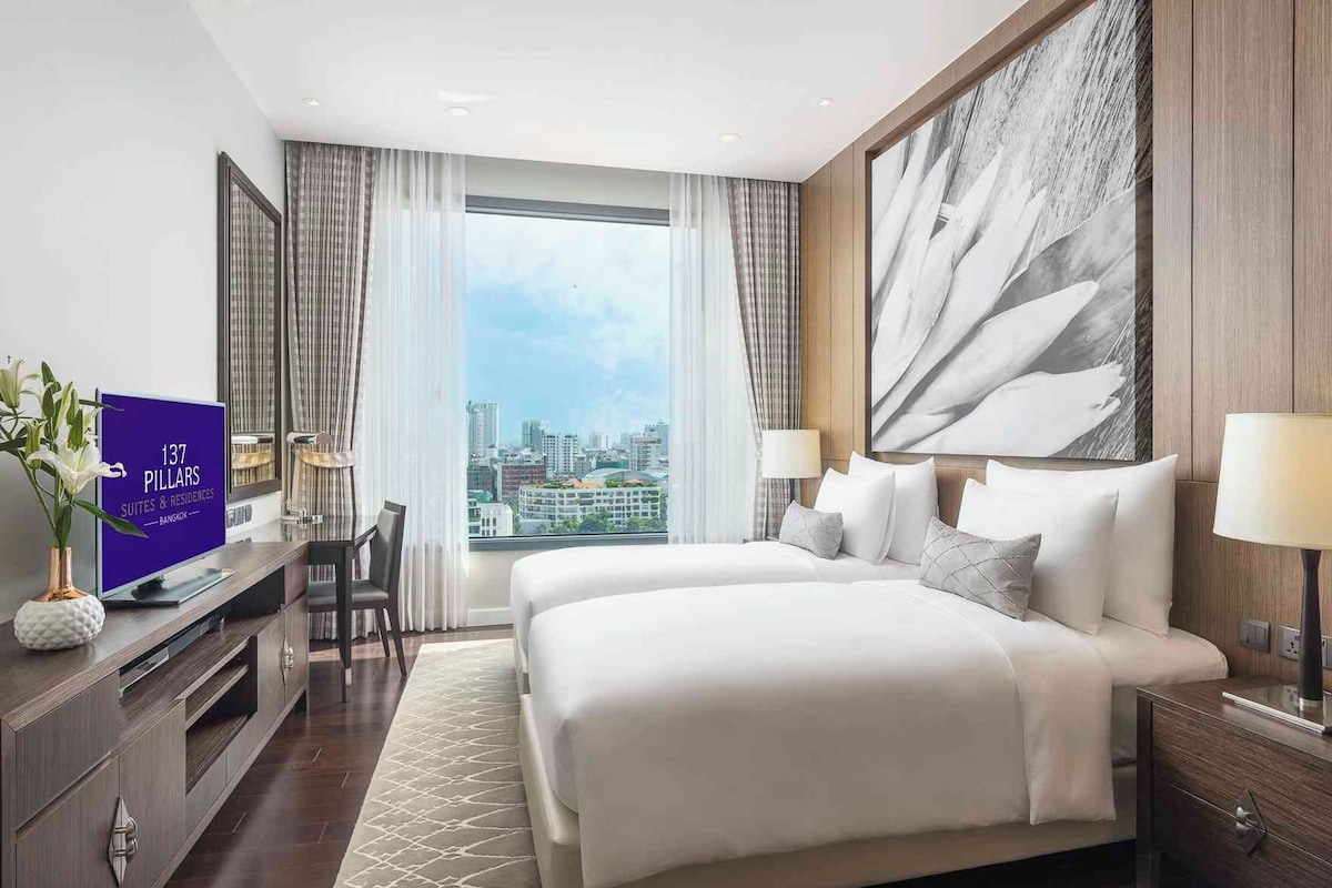 Een hotelkamer met een groot bed, een bureau met een computer, een televisie waarop kamerinformatie wordt getoond en een uitzicht op de stad.