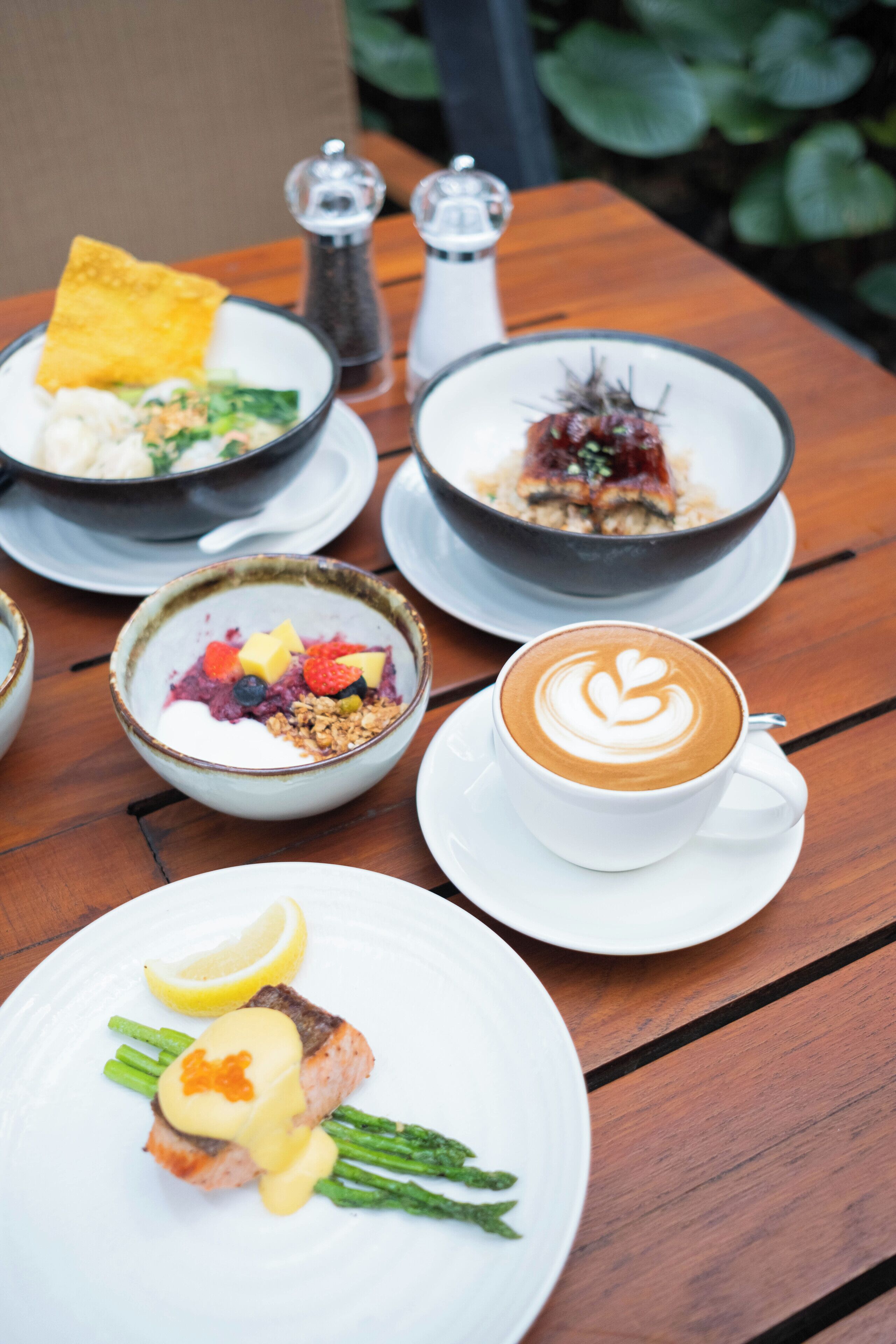 A table with various dishes including a bowl of soup, a plate with salmon and asparagus, a bowl of fruit salad, a cup of coffee with latte art, and a bowl of rice with fish.