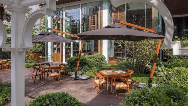 Une terrasse extérieure avec des tables et des chaises en bois, entourée d’une végétation luxuriante et avec un bâtiment en verre en arrière-plan.