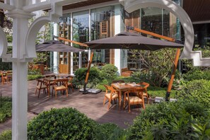 Une terrasse extérieure avec des tables et des chaises en bois, entourée d’une végétation luxuriante et avec un bâtiment en verre en arrière-plan.