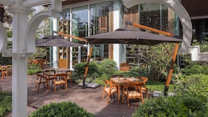 Een terras buiten met houten tafels en stoelen, omringd door weelderige begroeiing en een glazen gebouw op de achtergrond.