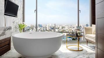 A modern bathroom with a large freestanding bathtub, a marble floor, a city view, a mounted TV, and a small table with a vase and flowers.