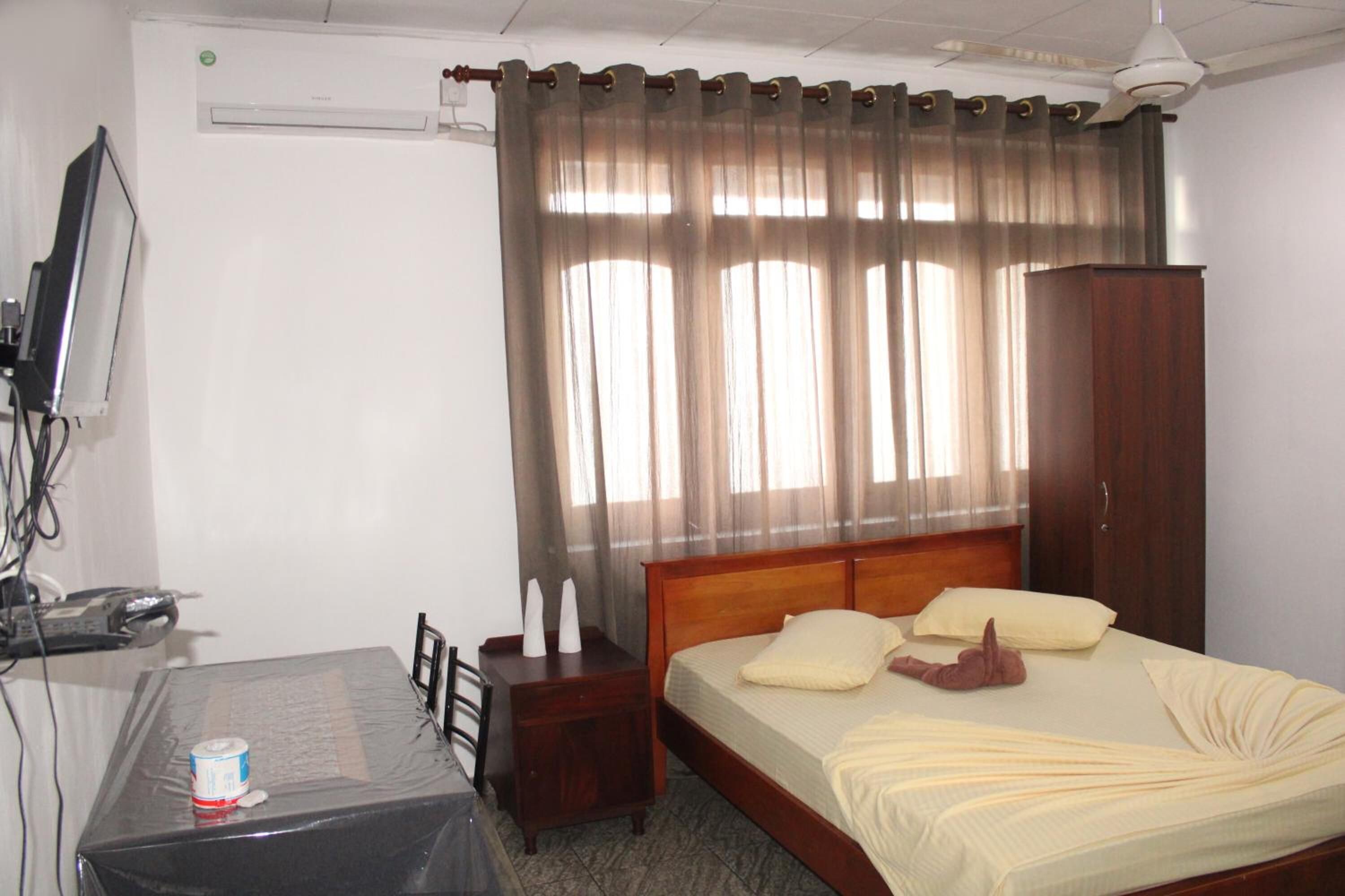 A hotel room with a bed, a wooden nightstand, a desk with a chair, a television, and a wardrobe.