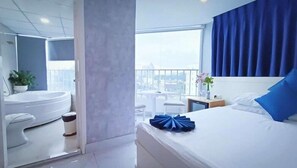 Ett modernt hotellrum med en säng, ett litet bord, en stol, ett fönster med utsikt över staden och ett badrum med badkar och handfat.
