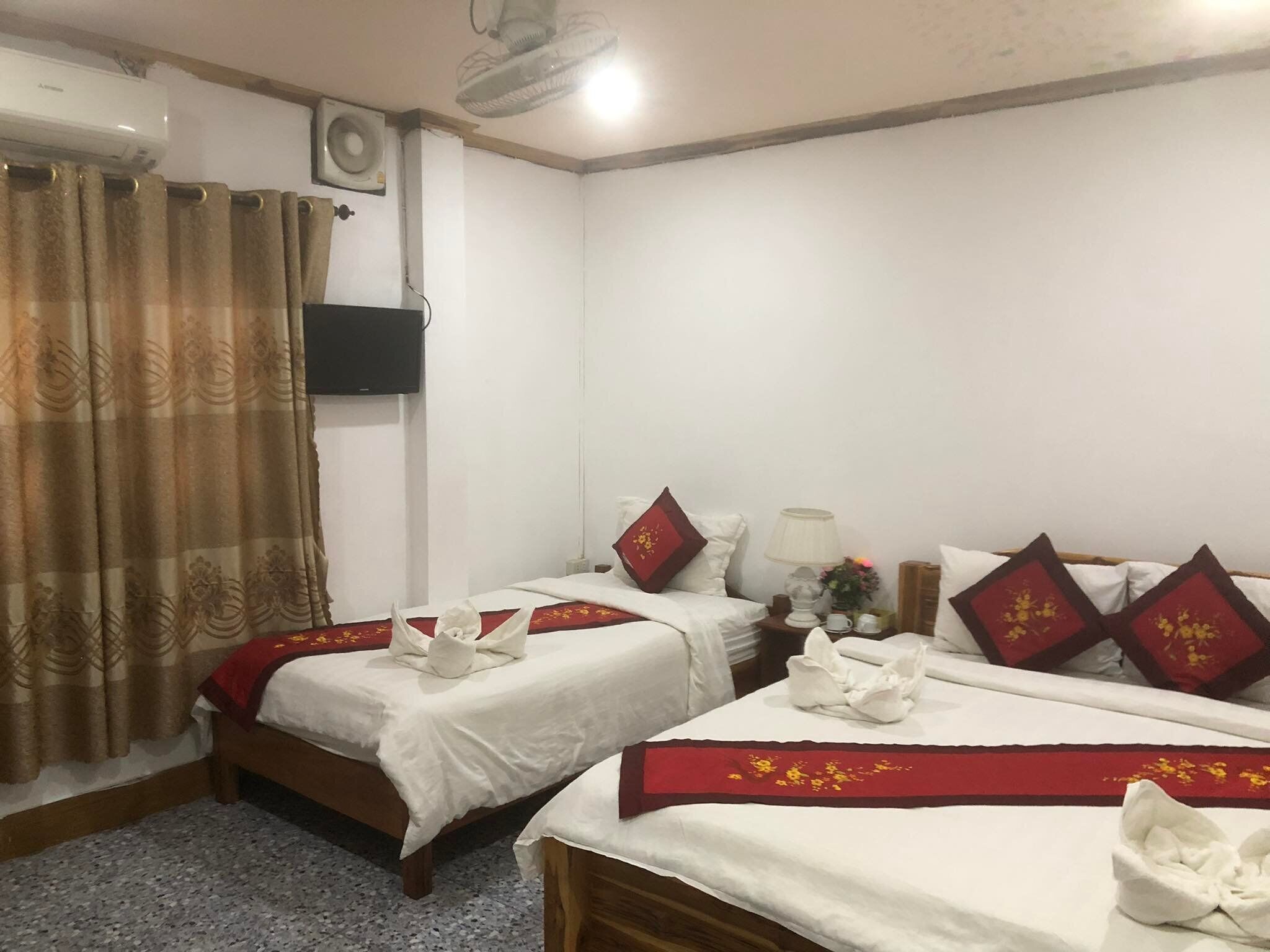 A hotel room with two beds, each with a red and white bedspread, white pillows, and a red decorative pillow. There is a ceiling fan, a wall-mounted air conditioner, and a TV on the wall.