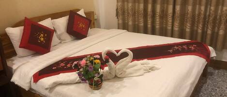 A neatly made bed with white linens, a red and white decorative pillow, a heart-shaped towel arrangement, and a small vase with flowers.