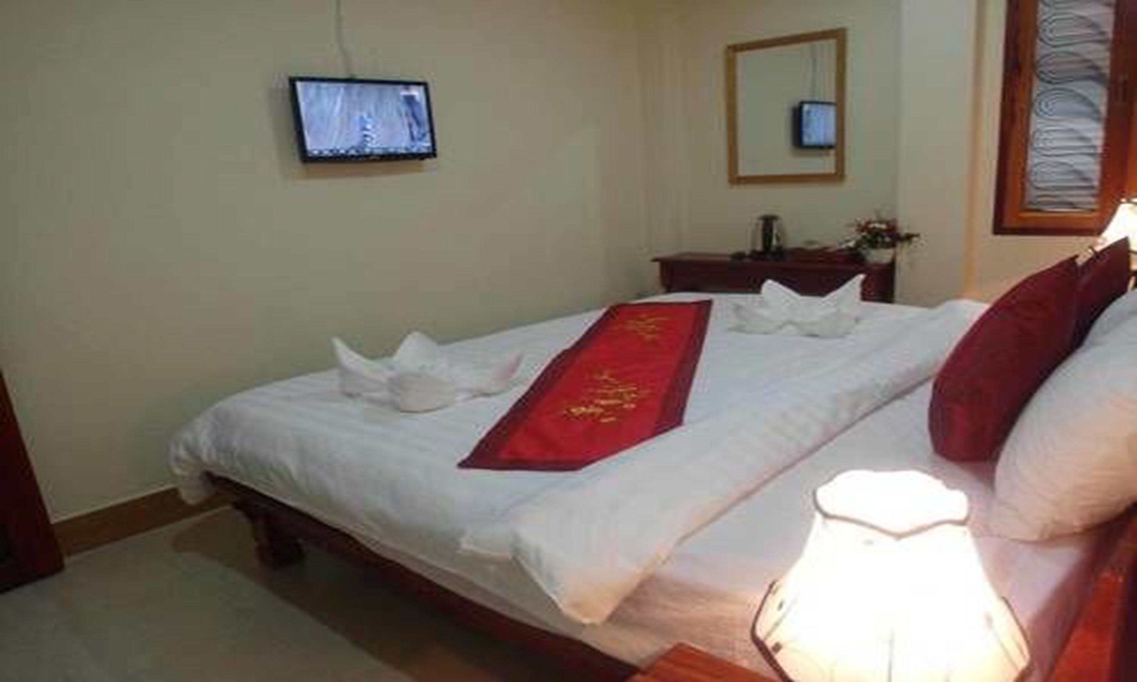 Un lit bien fait, agrémenté d’un coussin décoratif rouge et d’un coussin blanc. Un téléviseur est fixé au mur, à gauche du lit.