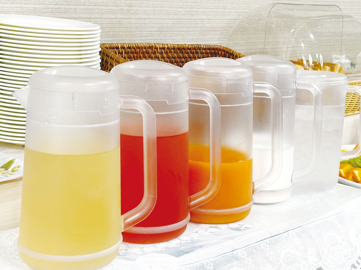 Una mesa con cuatro recipientes de vidrio con bebidas de colores distintos, una pila de platos blancos y una cesta de mimbre al fondo.
