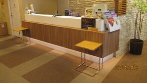 A reception desk with a wooden counter, two metal stools, a potted plant, and a signboard. - Hotel Green With (Sendai)