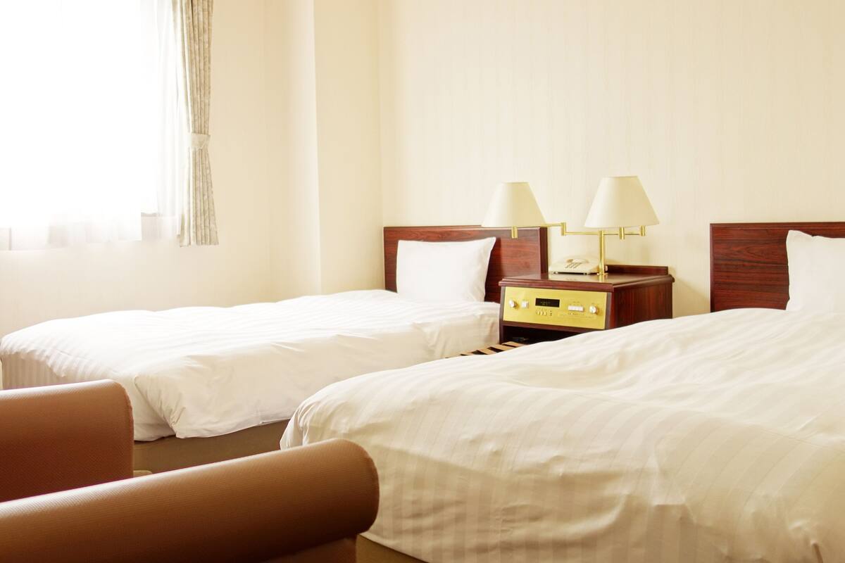 A hotel room with two single beds, a wooden nightstand with lamps, and a window with curtains.
