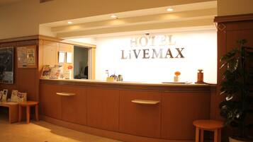 A reception desk with a sign reading "HOTEL LIVEMAX". There are wooden cabinets, a small stool, and a potted plant visible.