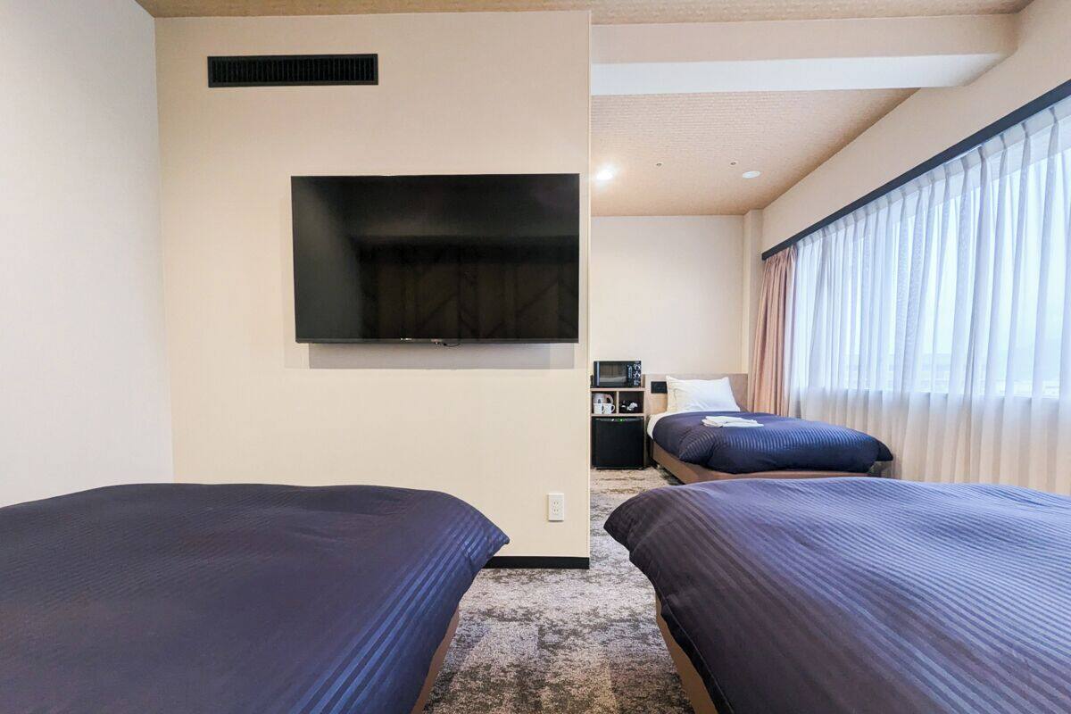 A hotel room with two beds, a TV mounted on the wall, a microwave, and a small kitchenette.