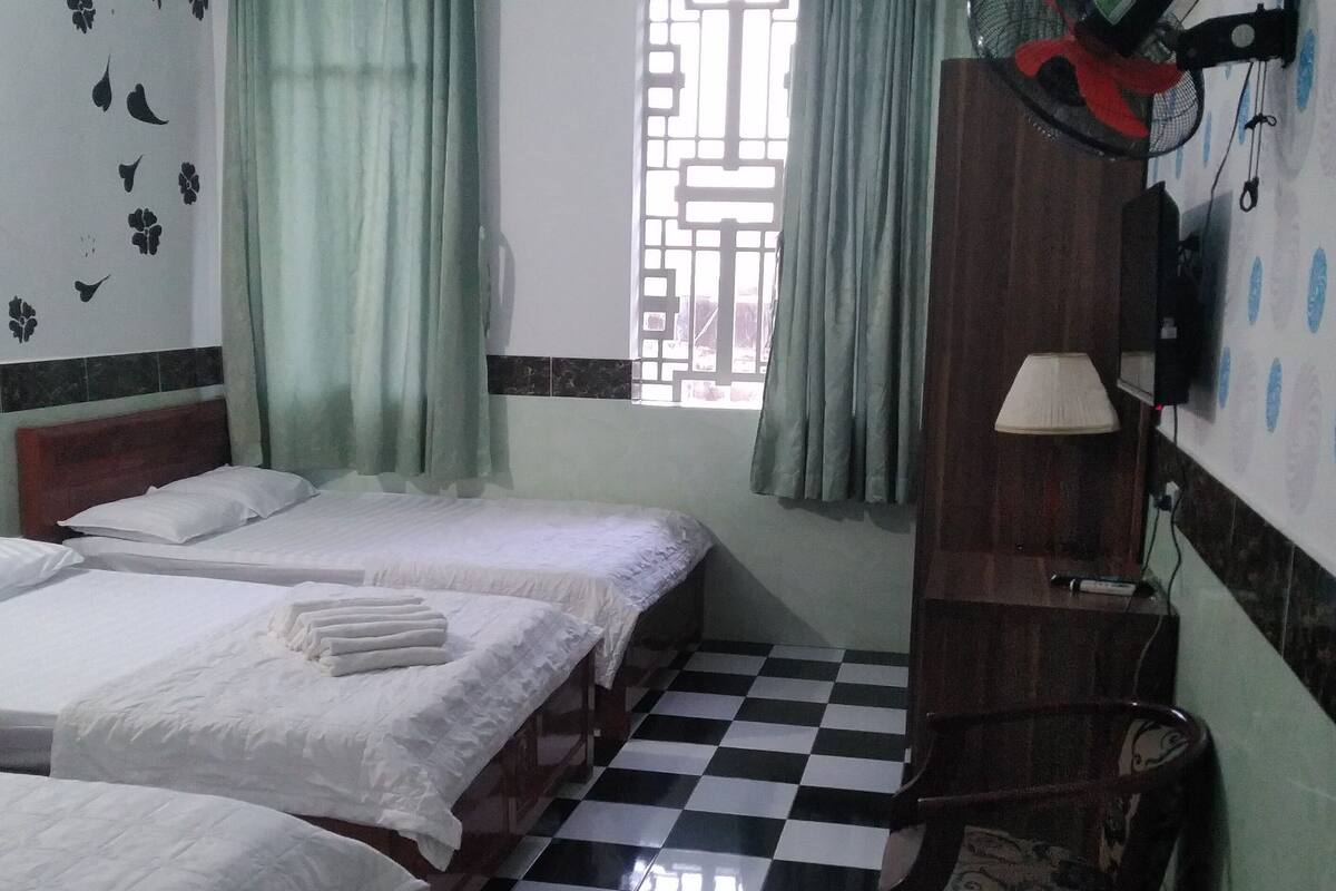 Habitació d'hotel amb tres llits, un sòl de quadres, una finestra amb cortines, un ventilador de sostre i una làmpada penjada a la paret.