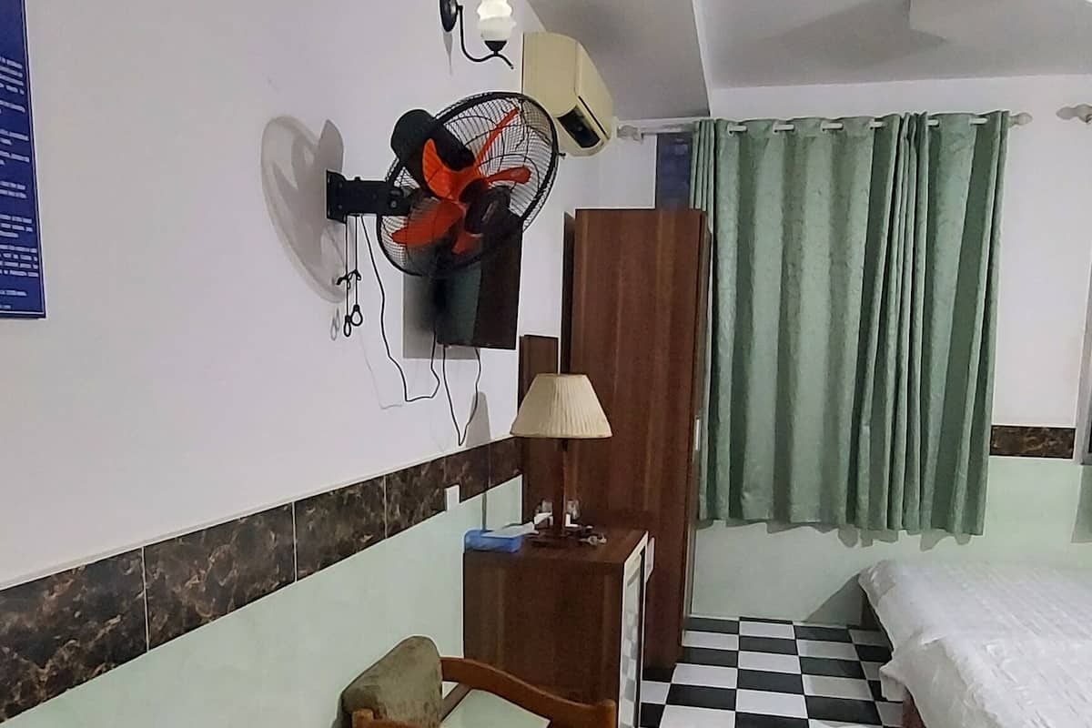 Habitació d'hotel amb un terra de pissos, un llit, una taula de nit amb una làmpada, una cadira, un ventilador fixat a la paret i una cortina verda.