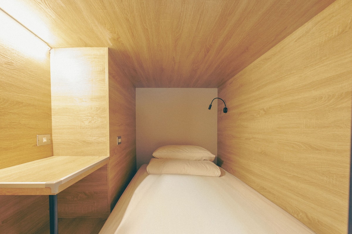 A small, wooden room with a single bed, a desk, and a wall-mounted lamp.