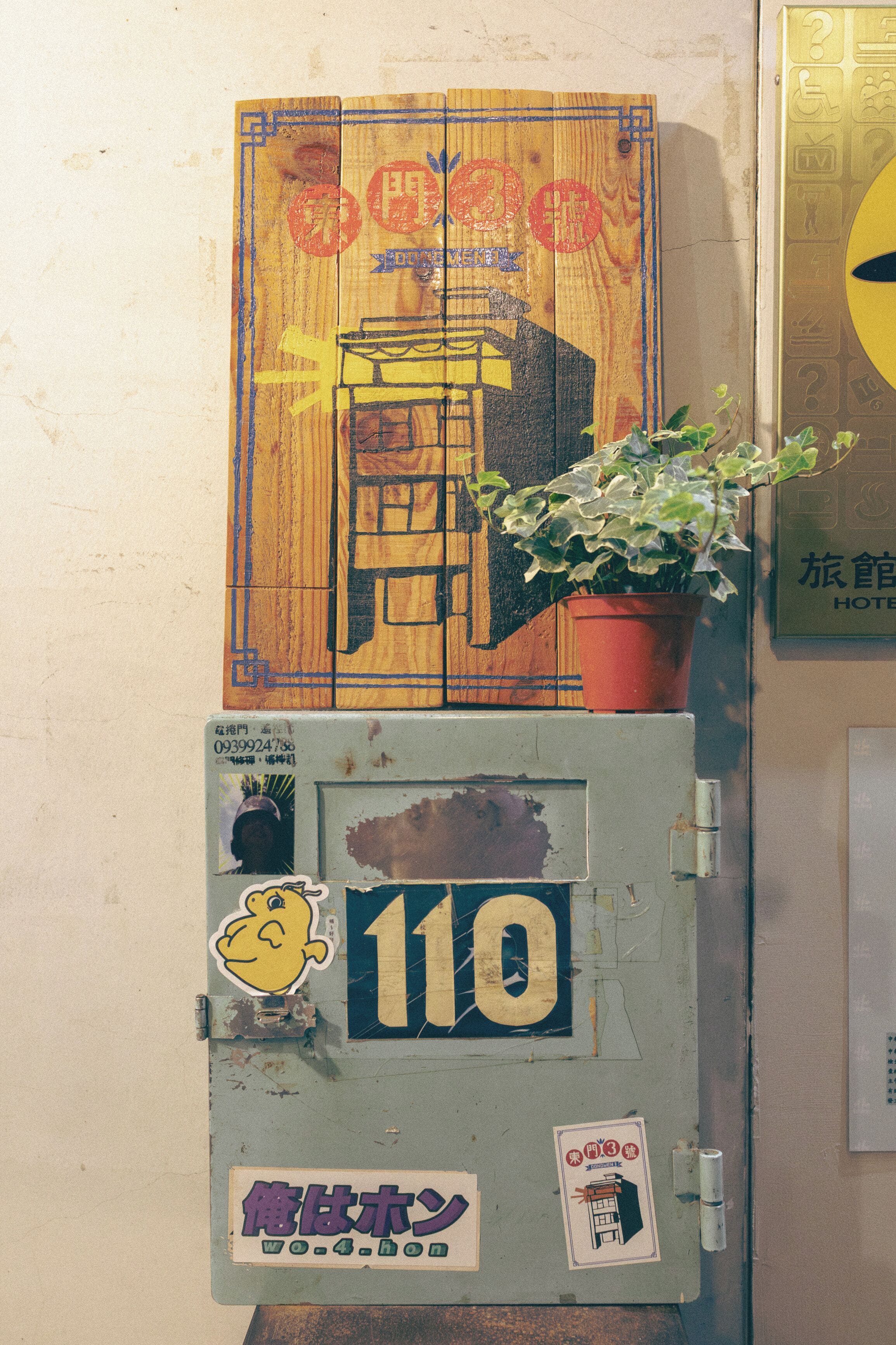A wall with a wooden panel, a potted plant, and a metal cabinet with stickers.