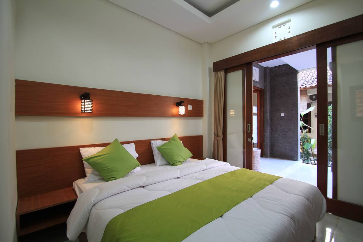 A hotel room with a wooden headboard, a large bed with white bedding and green accents, and a view of the outside through a glass door.